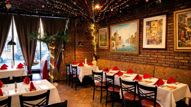 Cozy restaurant interior with brick walls, framed art, white tablecloths, red napkins, and a young girl seated.