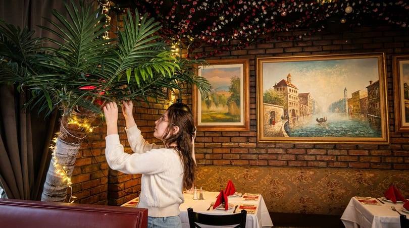 Woman decorating a palm tree with string lights in a restaurant with brick walls and paintings.