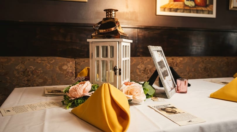 A white lantern centerpiece with candles, pink flowers, and yellow napkins on a white tablecloth.